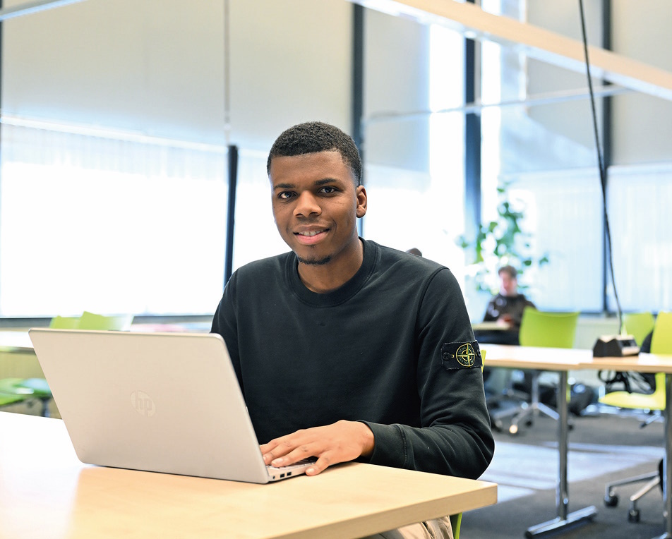 Studentenverhalen Jongen met zwarte trui in aan tafel met laptop, op de achtergrond zitten mensen.