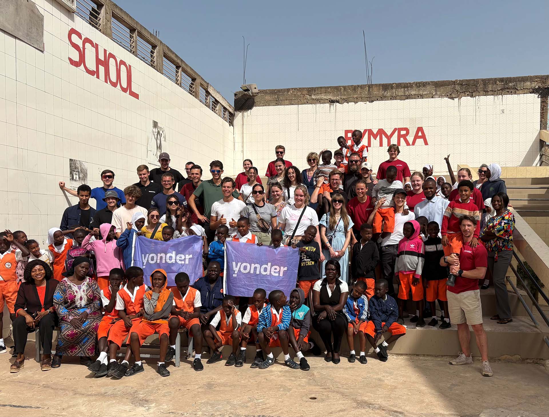 Yonder studenten werken samen tijdens de Excellentie reis naar Gambia Grote groep mensen, waaronder kinderen in uniform en volwassenen, poseert buiten voor een witte betegelde muur.